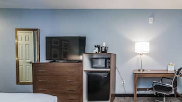 A hotel room with a bed, a wooden dresser, a microwave, a refrigerator, a desk with a lamp, and a television.