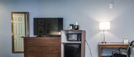 A hotel room with a bed, a wooden dresser, a microwave, a refrigerator, a desk with a lamp, and a television.