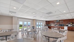 A spacious indoor dining area with multiple tables and chairs, a kitchen area with wooden cabinets, and large windows offering a view of the outdoors. - Ocean Isle Inn (Ocean Isle Beach)