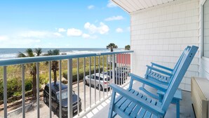 A balcony with a view of the ocean, a blue chair, and parked cars.