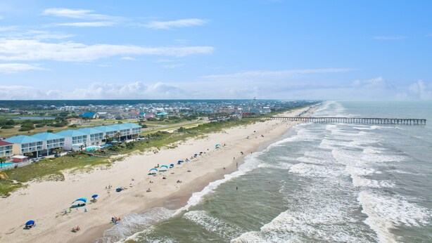 A beach with a pier extending into the ocean, a row of beachside buildings, and a sandy beach with umbrellas.