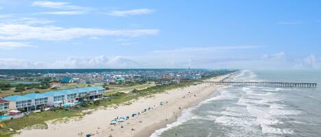 A beach with a pier extending into the ocean, a row of beachside buildings, and a sandy beach with umbrellas.