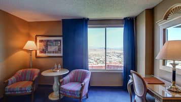 A hotel room with two striped armchairs, a small round table with a lamp, a desk with a lamp, and a large window offering a city view.
