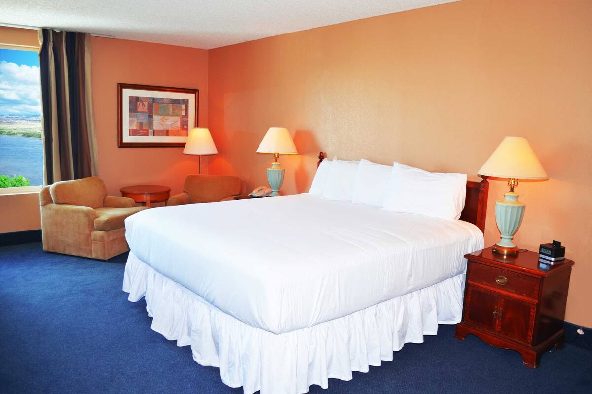 A hotel room with a large bed, two bedside lamps, a chair, a small table, and a window with a view of the outdoors.