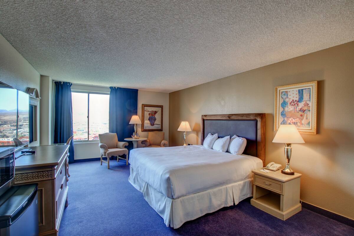 A hotel room with a large bed, a desk with a microwave, two chairs, a nightstand with a lamp, and a painting on the wall.
