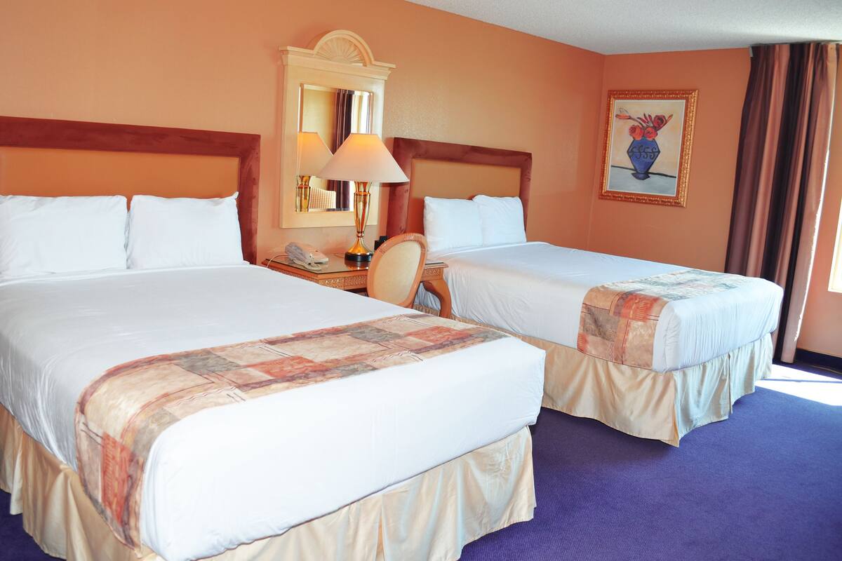 A hotel room with two beds, a nightstand, a lamp, a chair, a mirror, and a painting on the wall.