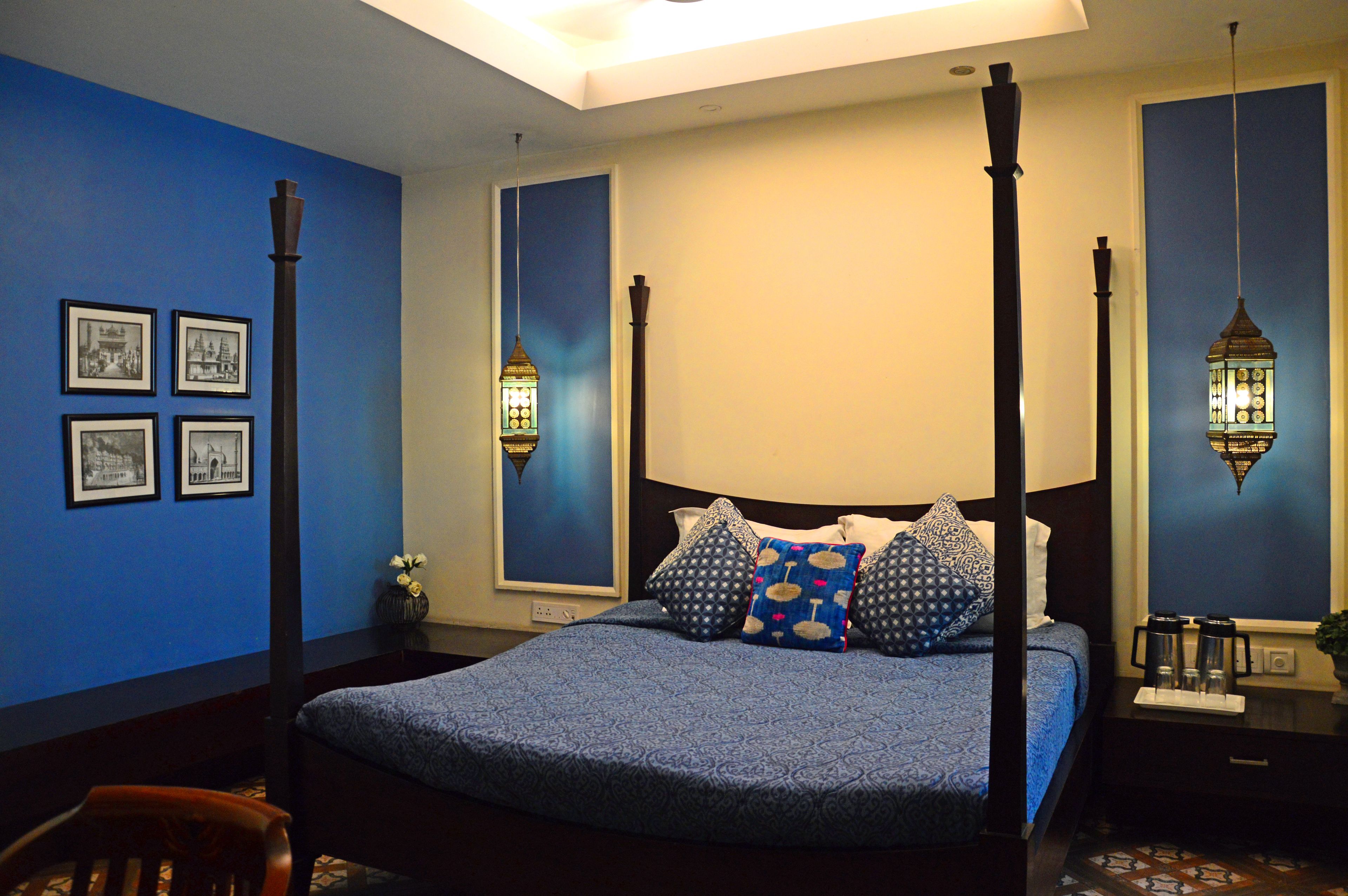 Uma cama com dossel com colcha azul e almofadas estampadas, mesa de cabeceira com abajur e quadros nas paredes.