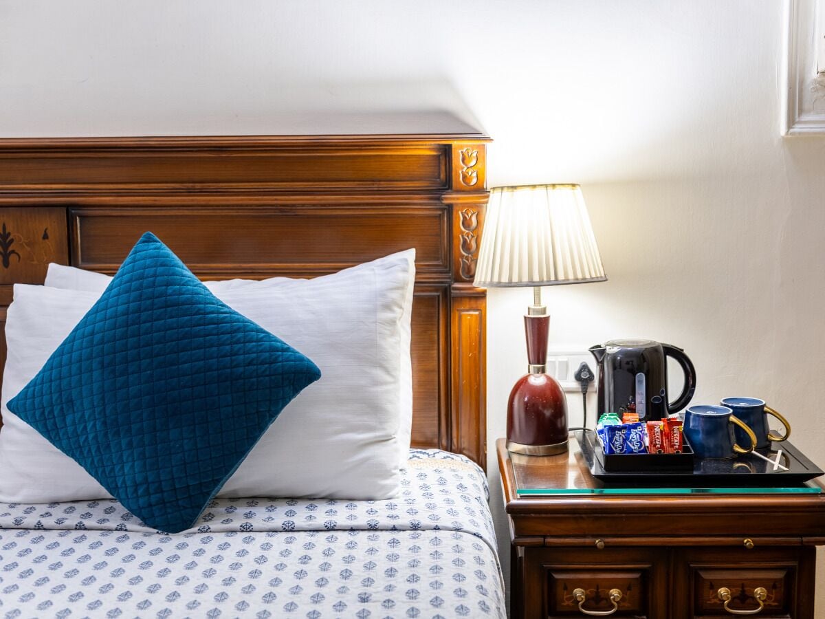 Una cama bien hecha con una colcha estampada y dos almohadas, una azul y otra blanca. Se ve una mesita de noche con una lámpara, una bandeja con tazas y una cafetera.