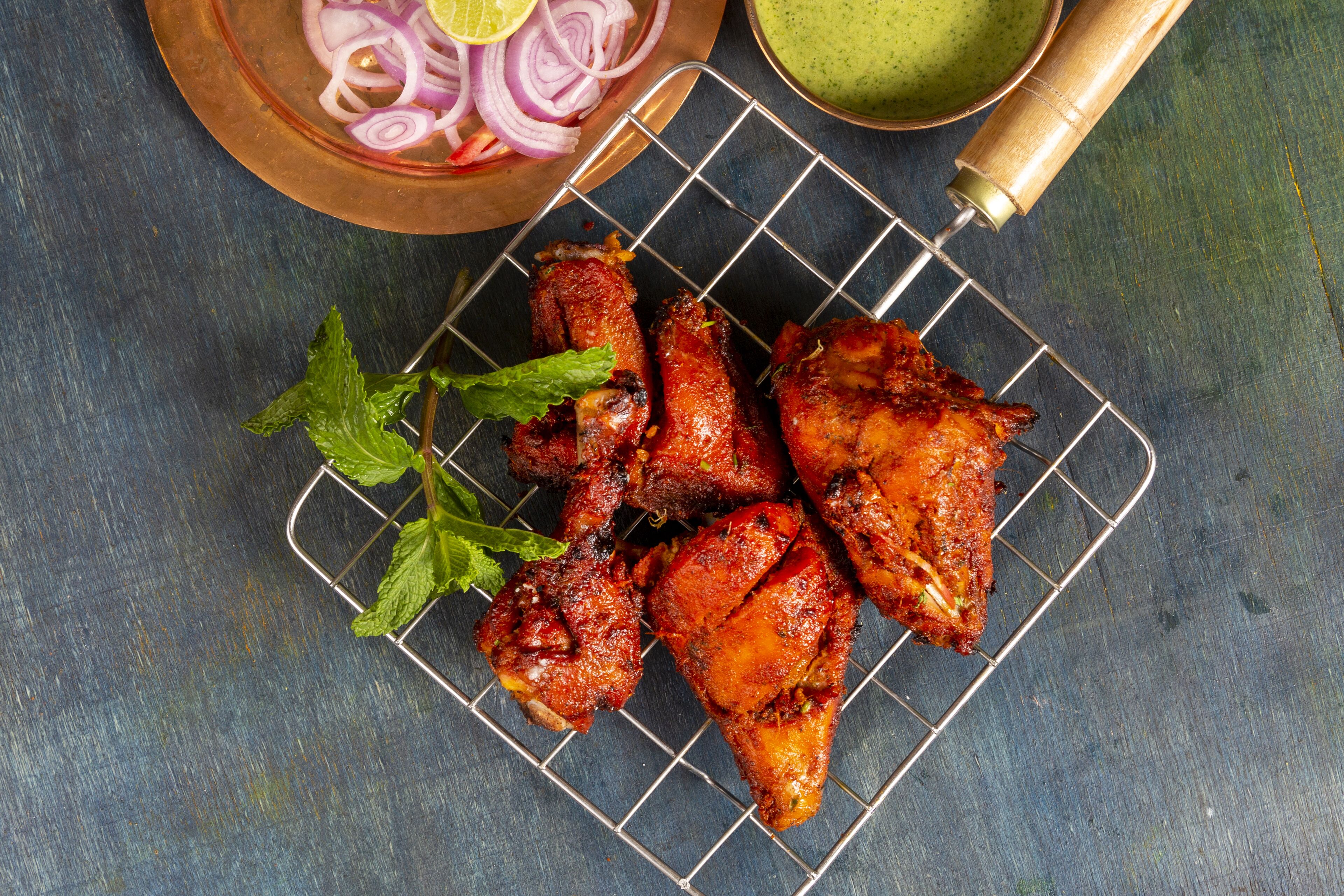 Grilled chicken wings on a wire rack with lemon slices and red onion rings, accompanied by a green sauce container.