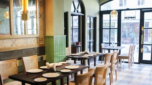 L’intérieur d’un restaurant présente de nombreuses tables et chaises, un sol à carreaux et de grandes fenêtres donnant sur la rue.