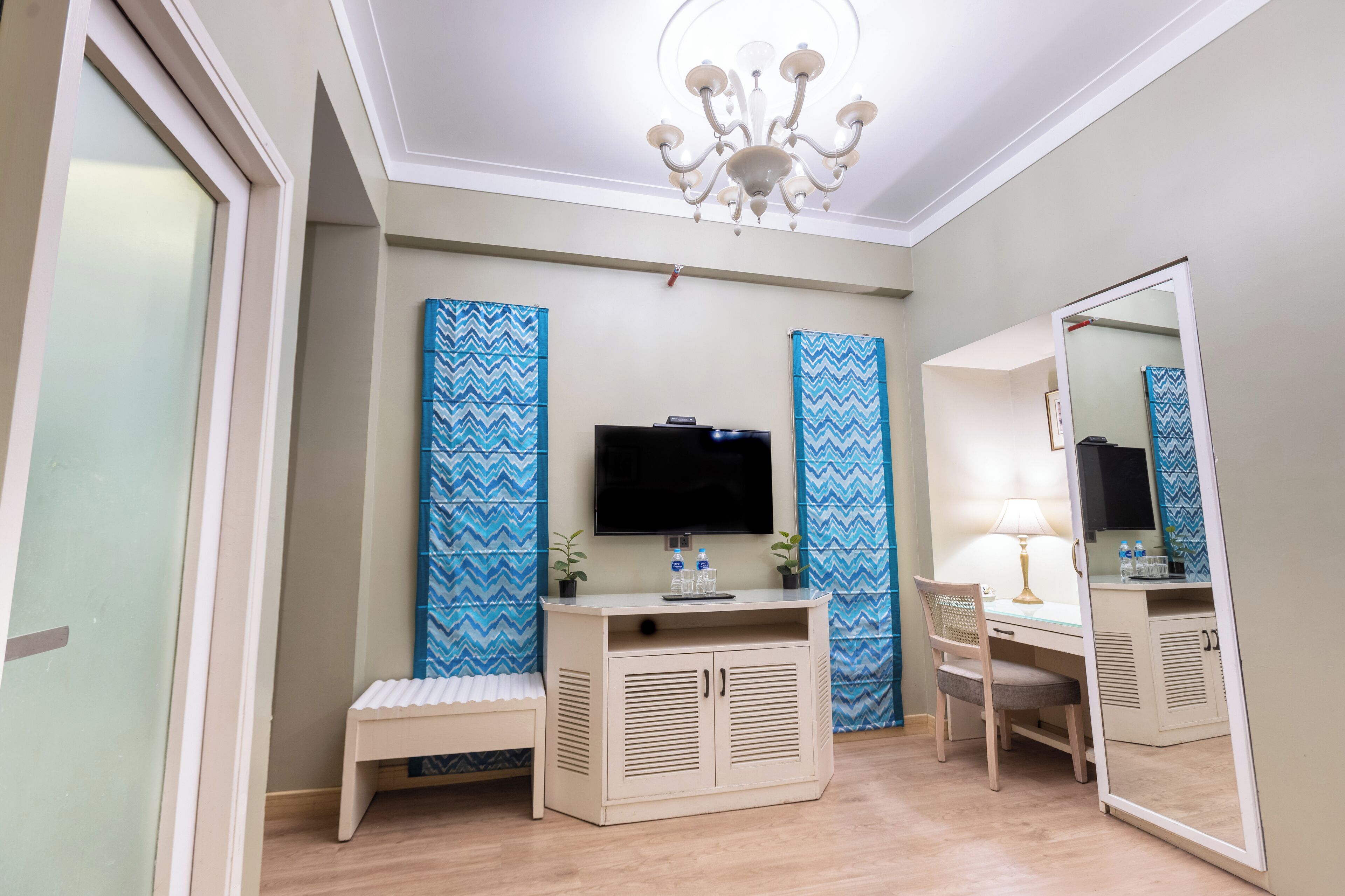 A bedroom with a TV, a desk, a chair, a bench, and a mirror.