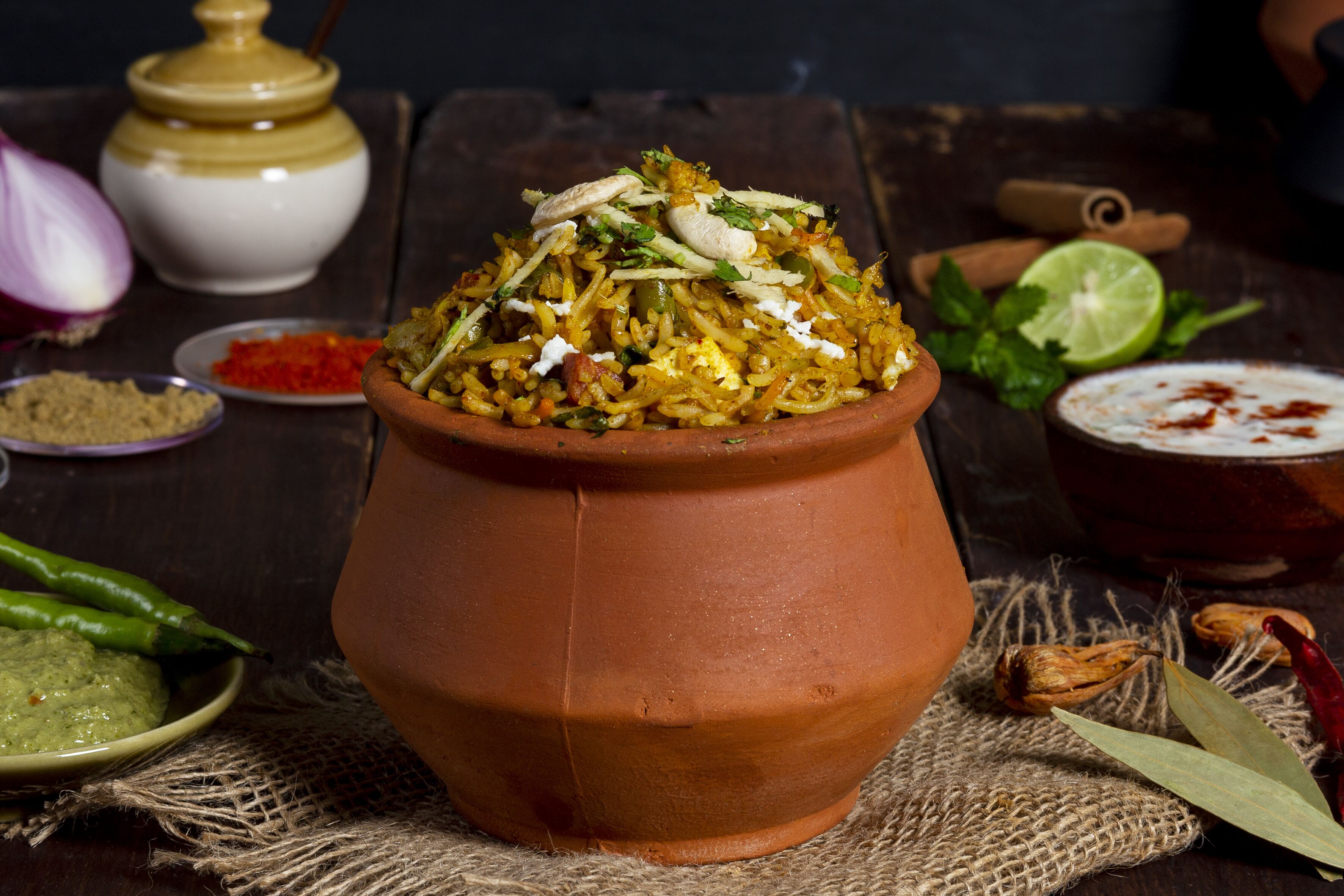 Una olla de barro llena de un plato de arroz adornado con hierbas y nueces, rodeada de especias y un limón en una mesa rústica.