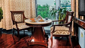 Un coin repas avec deux chaises en bois et une table ronde, offrant une vue sur un balcon agrémenté de verdure et, au loin, sur une enseigne d’hôtel.