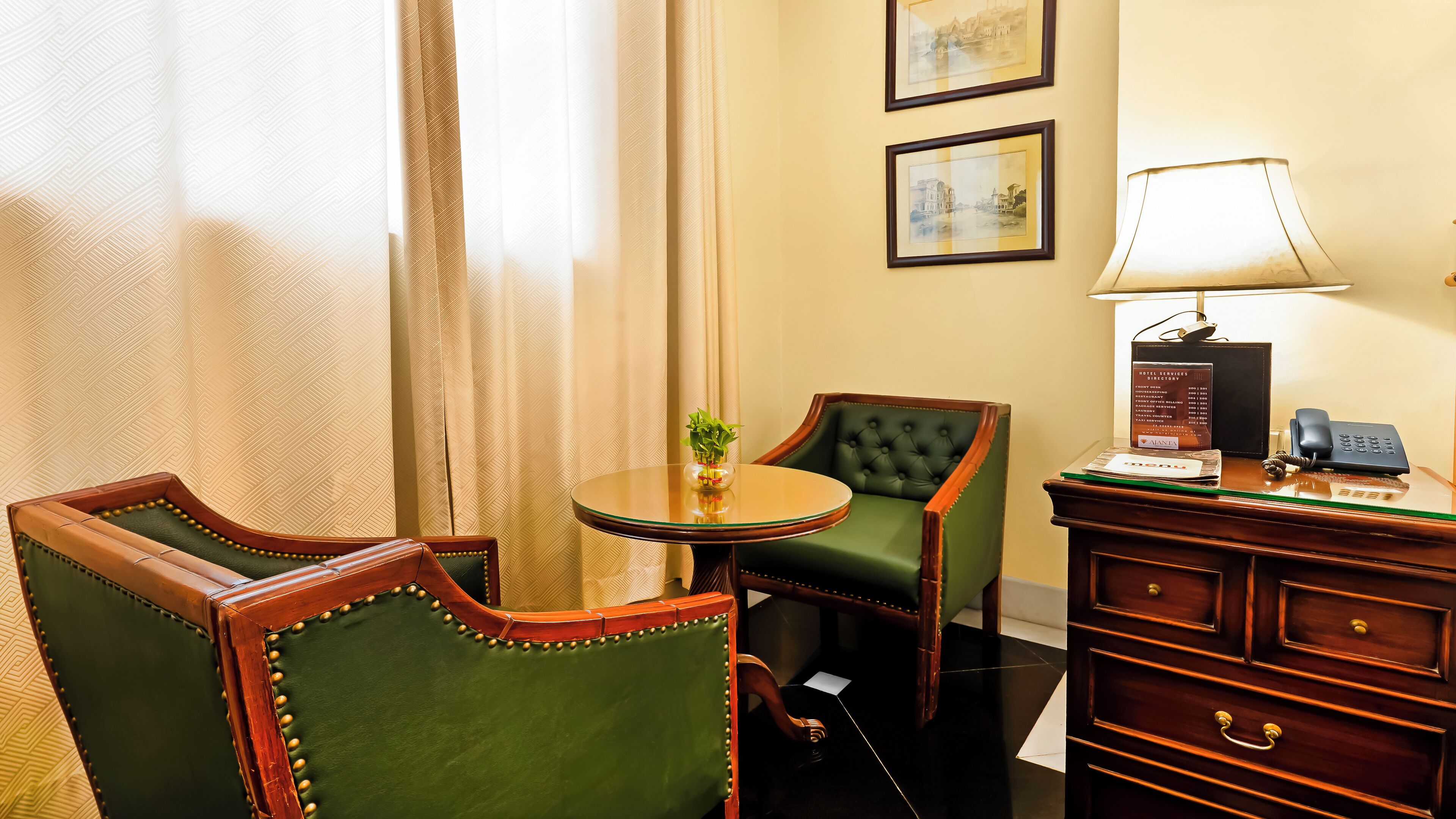 A hotel room with a green upholstered chair, a wooden chair, a small round table with a vase of flowers, a lamp, a telephone, and a framed menu on a wooden cabinet.