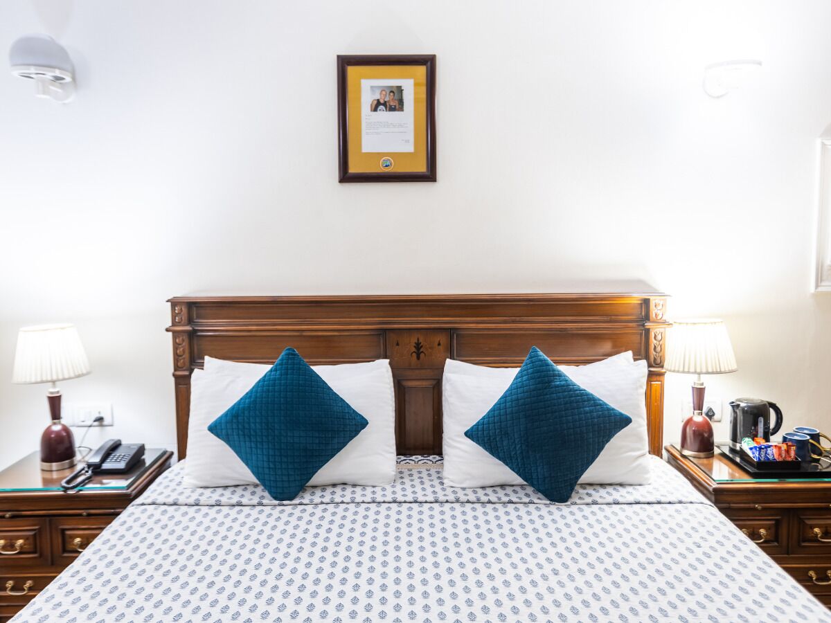 Habitación de hotel con cabecera de madera, dos almohadas azules, colcha estampada, un certificado enmarcado en la pared, una lámpara de noche, un teléfono y una bandeja con tazas de café y aperitivos.