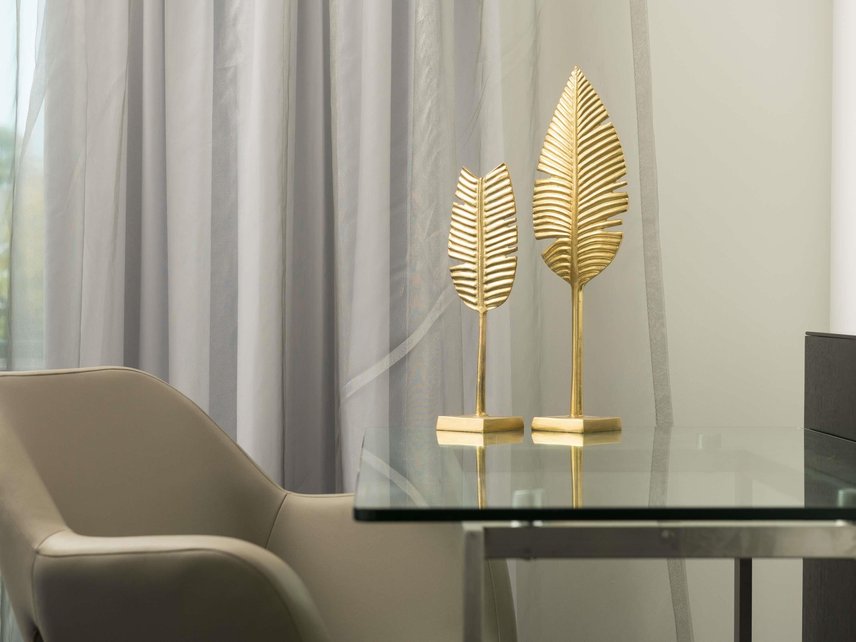 Una habitación moderna con una mesa de vidrio, dos adornos dorados con forma de hoja y una silla color beige.