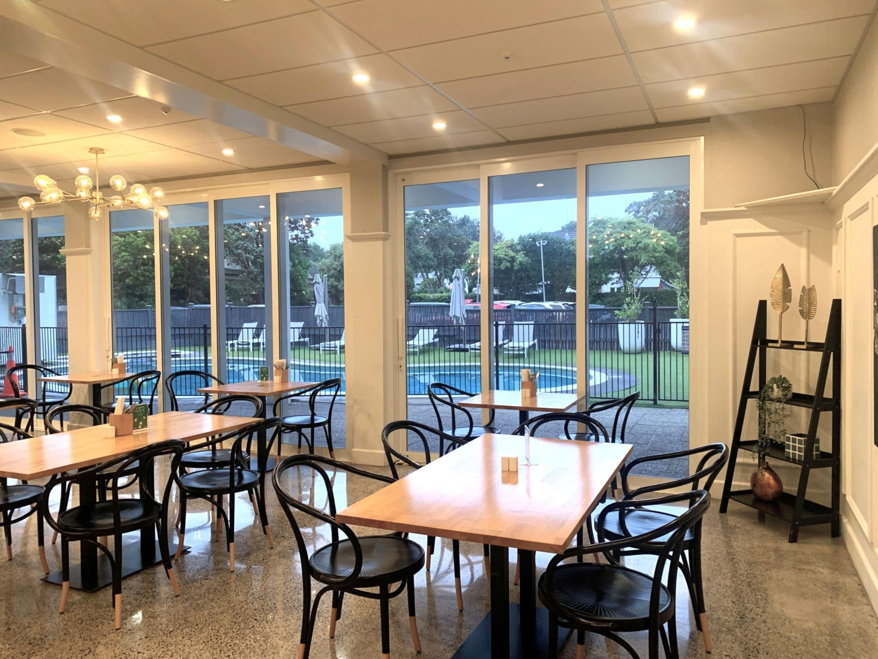 A restaurant with multiple tables and chairs, large windows offering a view of a swimming pool and outdoor seating.