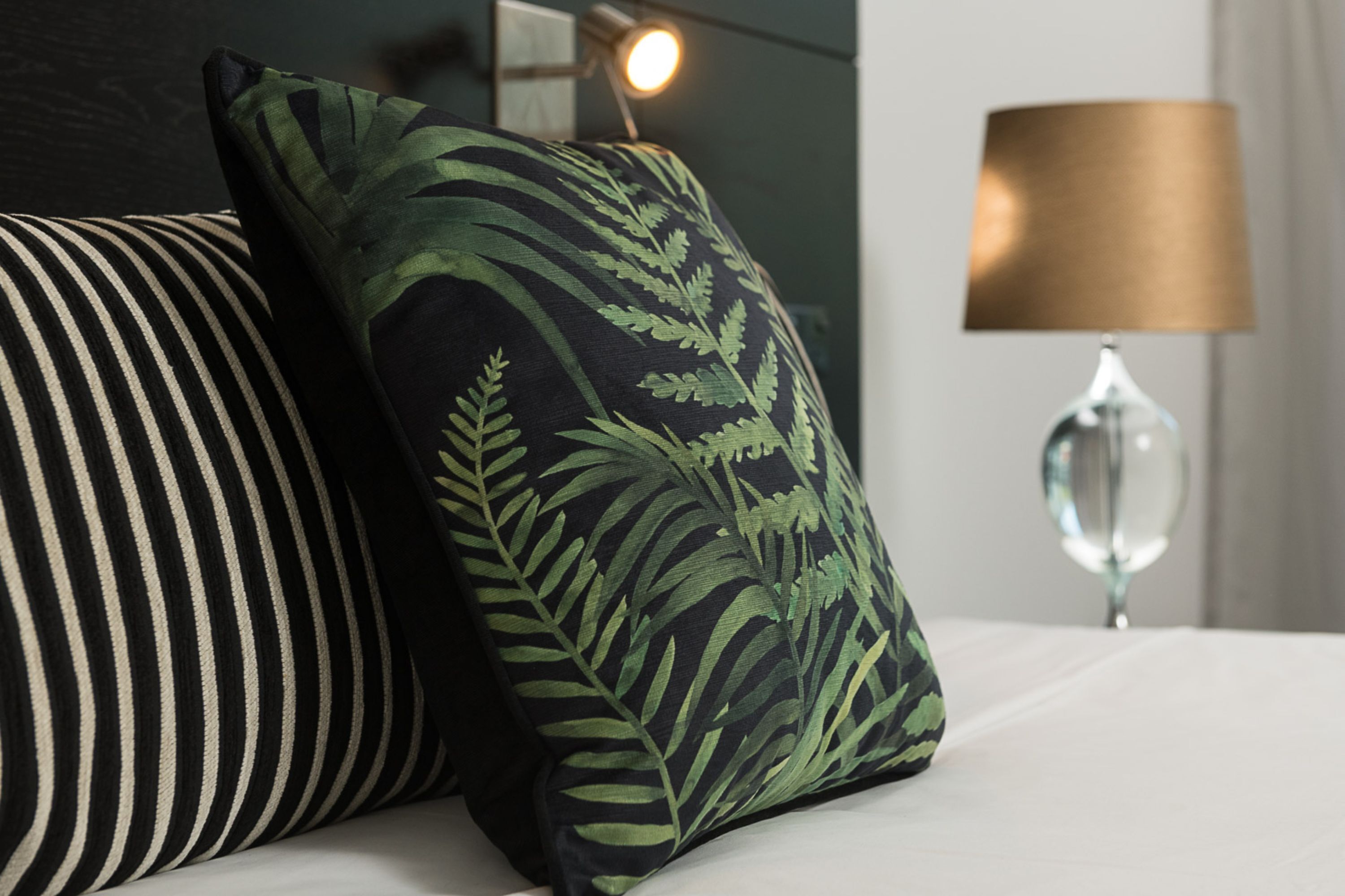 A bed with striped and fern-patterned pillows, a clear glass lamp, and a wall-mounted light.