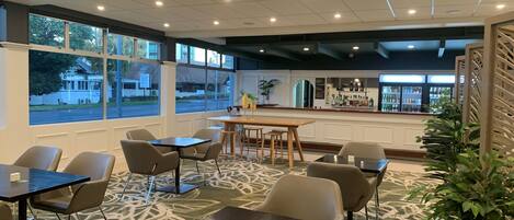 Un espace de restauration intérieur moderne, doté de nombreuses tables et chaises, de grandes fenêtres offrant une vue sur l’extérieur et d’un bar situé au fond de la pièce.