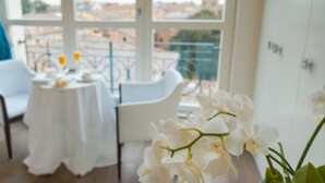 Uma sala de jantar com mesas e cadeiras brancas, uma varanda com guarda-corpo e flores brancas em primeiro plano.