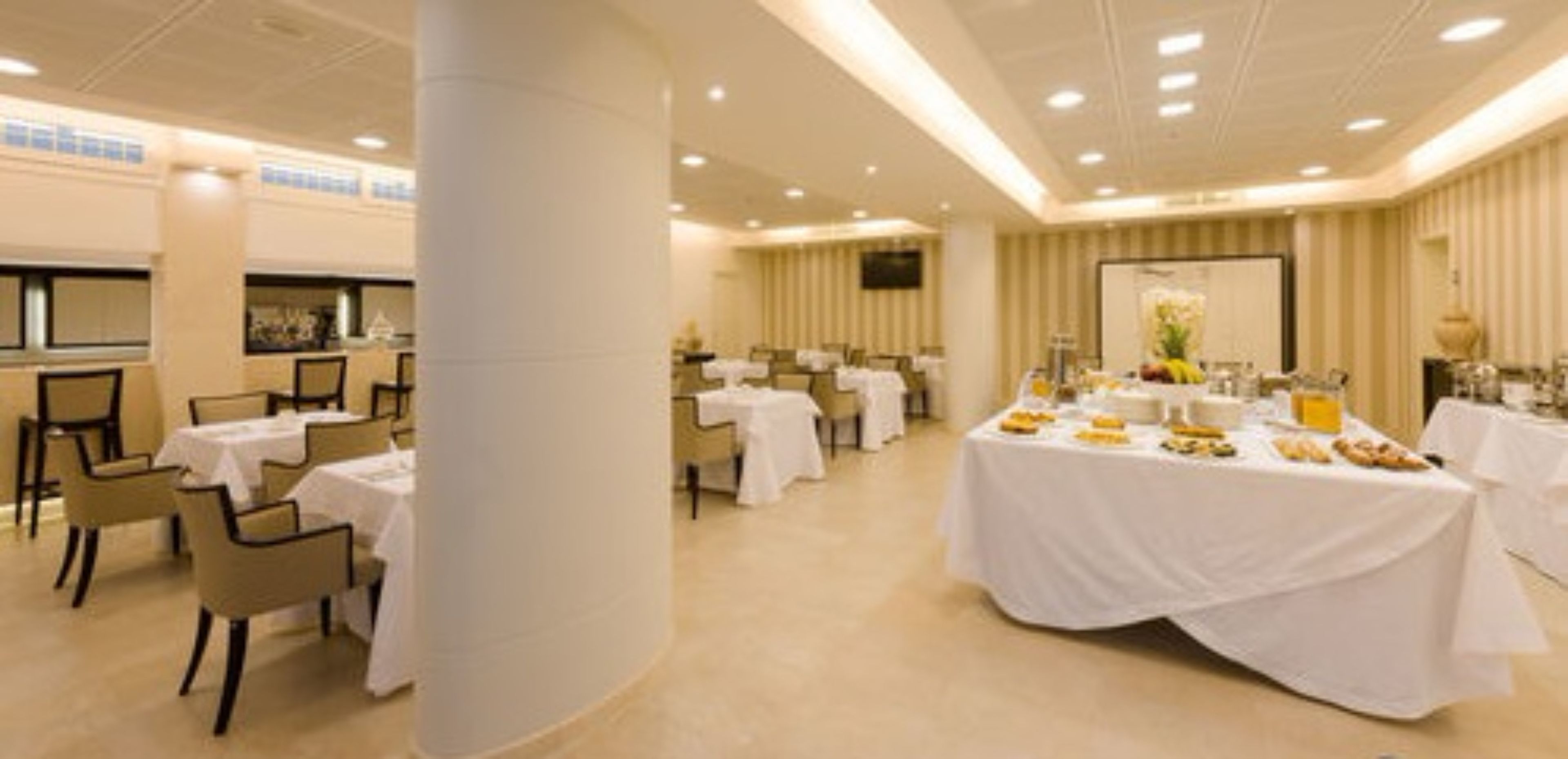A hotel dining area with round tables, white tablecloths, and a buffet setup.