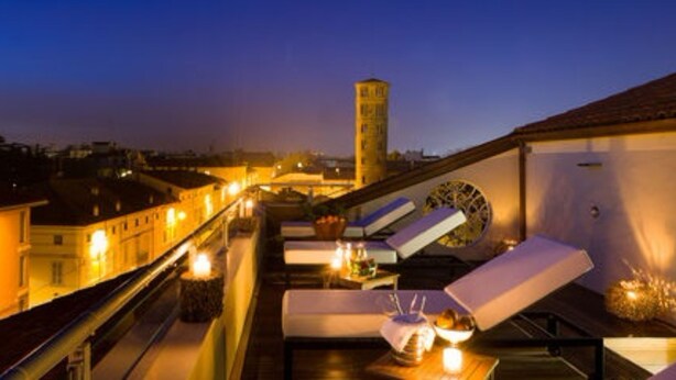 Terraço no telhado com vista para a cidade, incluindo uma torre. Há cadeiras de lounge e mesas com velas, sugerindo um espaço ao ar livre relaxante.
