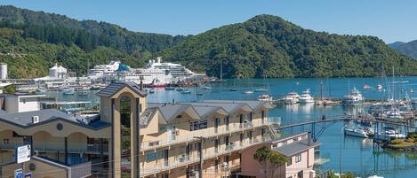 A coastal hotel with multiple balconies, a marina with boats, and a forested hill in the background.