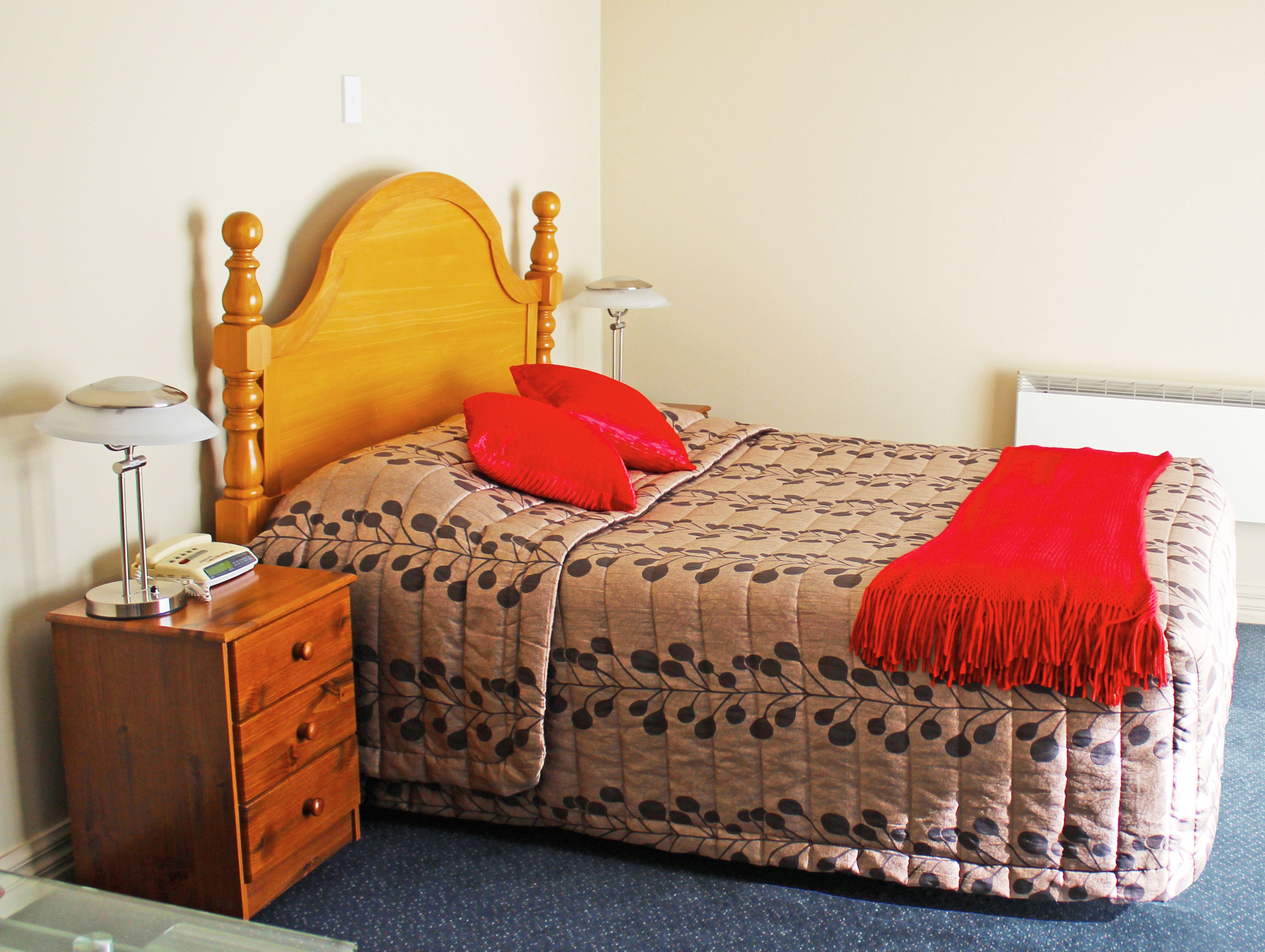 A neatly made bed with a floral-patterned duvet, two red pillows, and a red throw blanket. There is a wooden nightstand with a lamp and a telephone beside the bed.