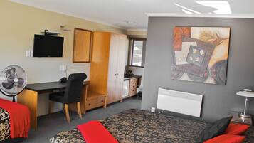 A compact hotel room with a bed, a desk, a chair, a TV, a fan, and a radiator.