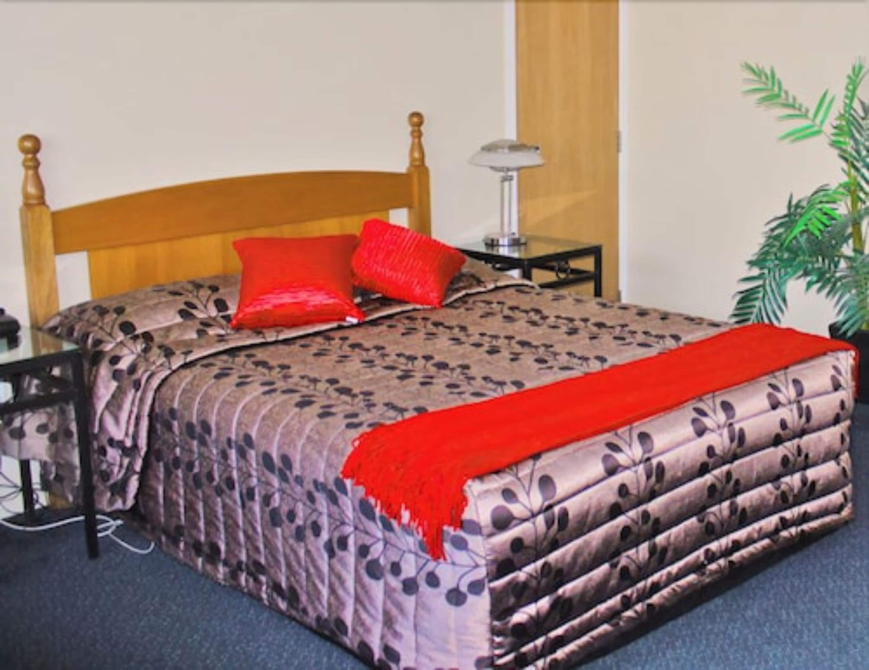 Una cama bien hecha con cubrecama estampado floral, dos almohadas rojas y una manta roja. Hay una mesita de noche con una lámpara y una pequeña planta en maceta.