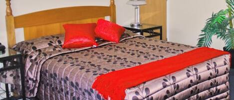 Una cama bien hecha con cubrecama estampado floral, dos almohadas rojas y una manta roja. Hay una mesita de noche con una lámpara y una pequeña planta en maceta.