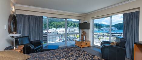 A hotel room with a large bed, two armchairs, a TV, and a view of a cityscape and water.