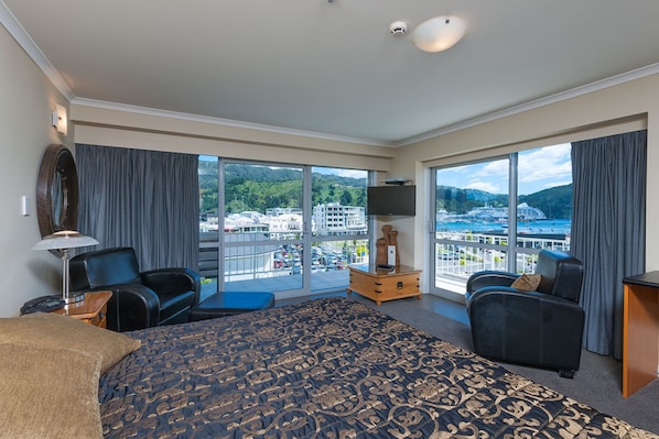 A hotel room with a large bed, two armchairs, a TV, and a view of a cityscape and water.