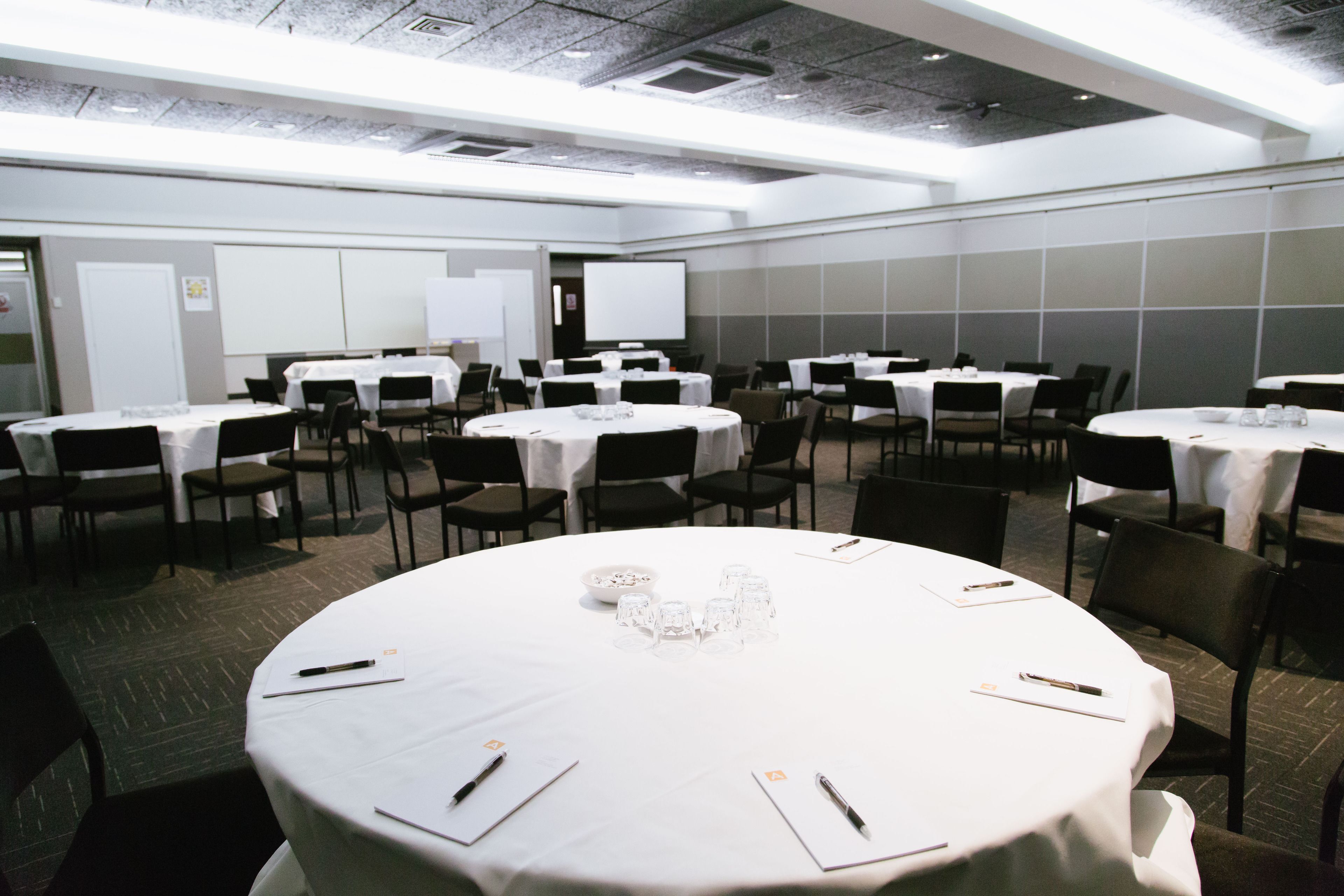 Une salle de conférence dotée de plusieurs tables rondes, chacune étant agrémentée de chaises, de nappes blanches et de stylos.