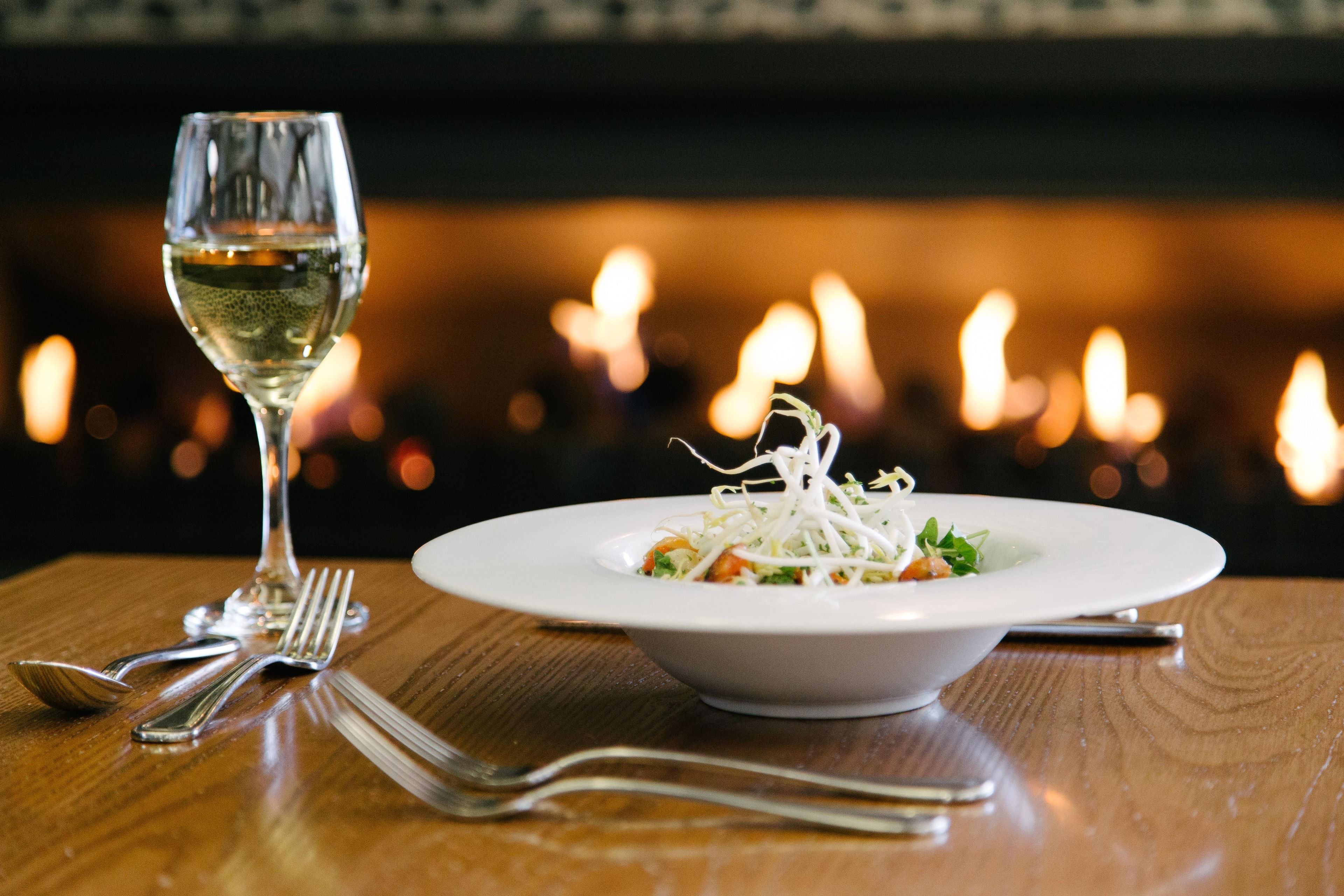 A dining table with a white plate of food, a glass of white wine, and cutlery.