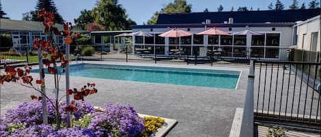 A swimming pool with a fenced deck area, surrounded by purple and yellow flowers, and outdoor seating with umbrellas.