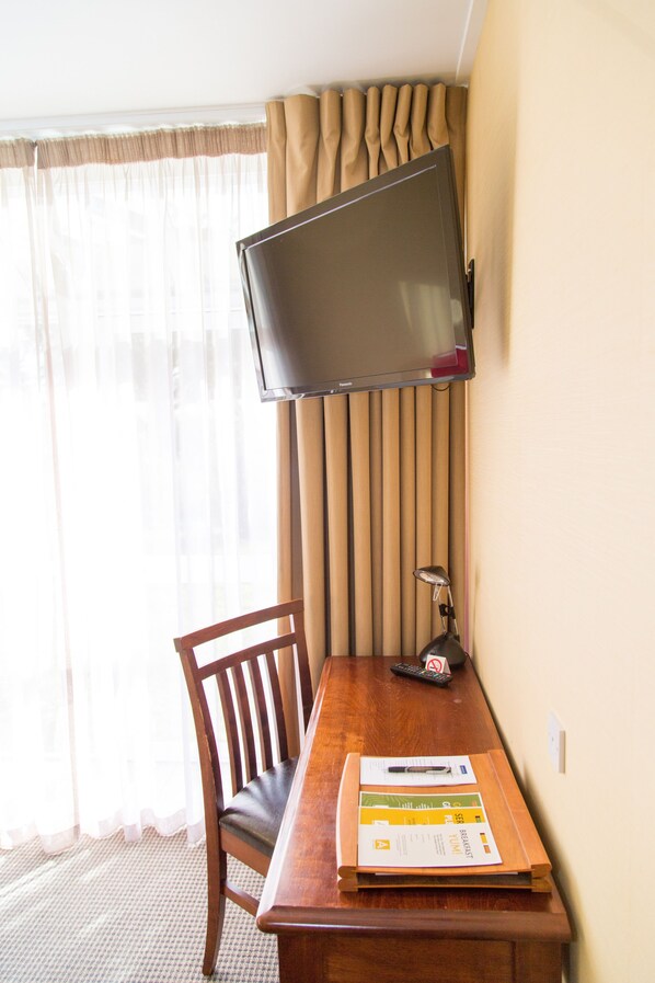 Habitación de hotel con un escritorio de madera, una silla, un televisor de pantalla plana montado en la pared y una ventana con cortinas translúcidas.