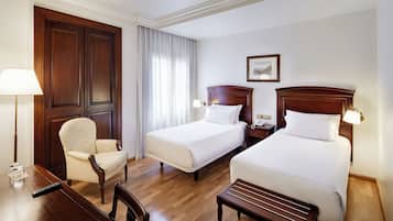 Habitación de hotel con dos camas individuales, un escritorio de madera, una silla, una lámpara y un cuadro en la pared.