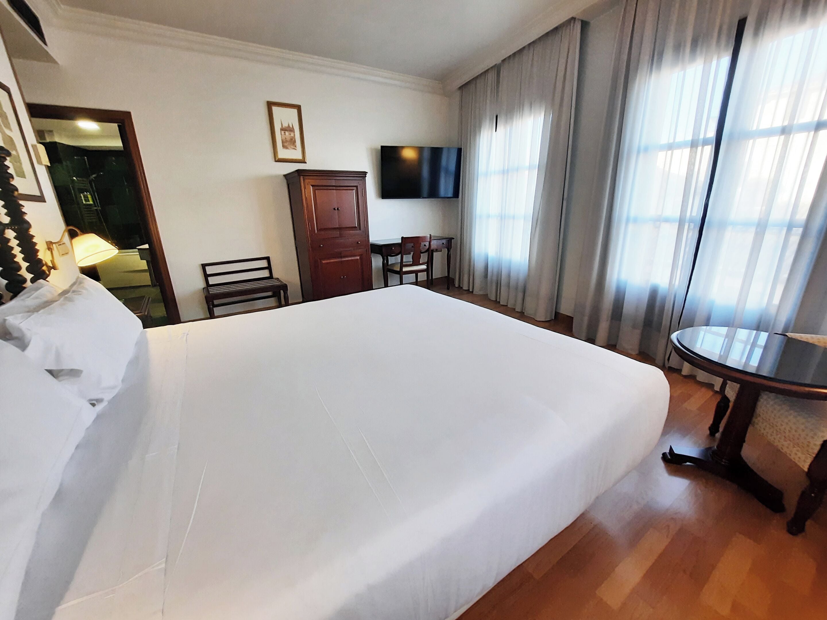 A hotel room with a large bed, a desk, a chair, a television, and a wardrobe.