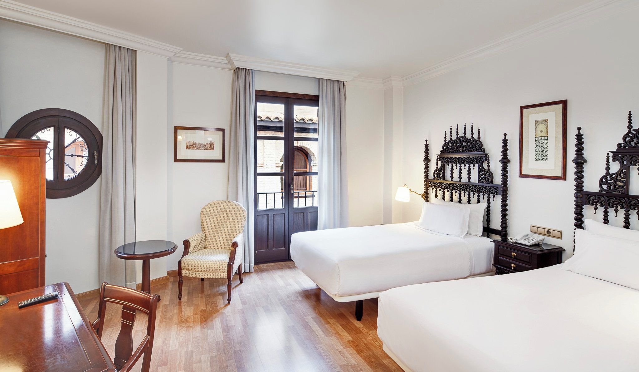 A hotel room with a bed, a chair, a small table, and a desk with a lamp.