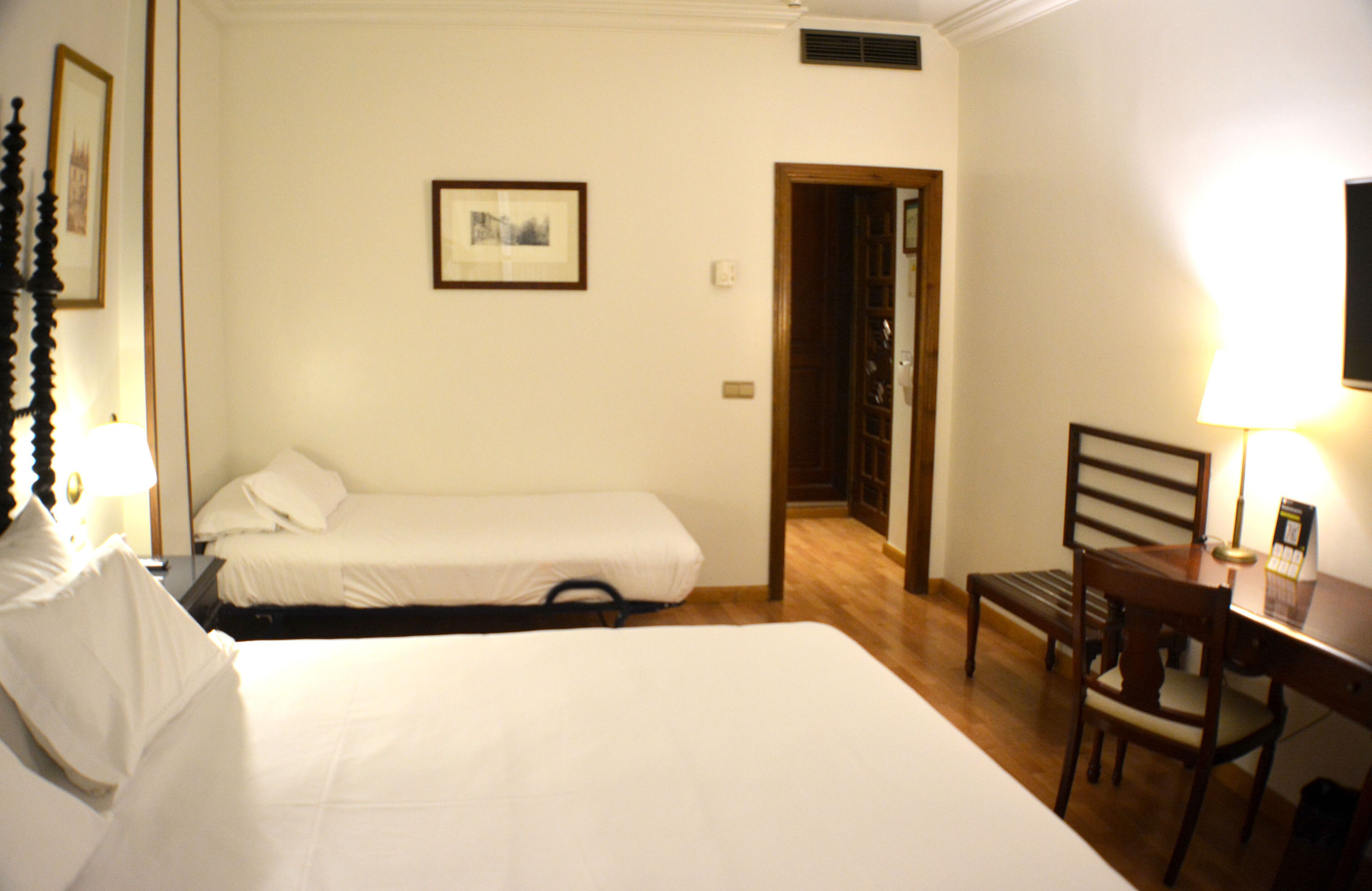 Habitación de hotel con dos camas individuales, un escritorio de madera con una silla, un cuadro enmarcado en la pared y una puerta que da a otra habitación.