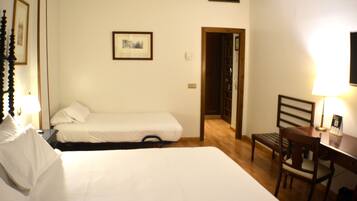 Habitación de hotel con dos camas individuales, un escritorio de madera con una silla, un cuadro enmarcado en la pared y una puerta que da a otra habitación.