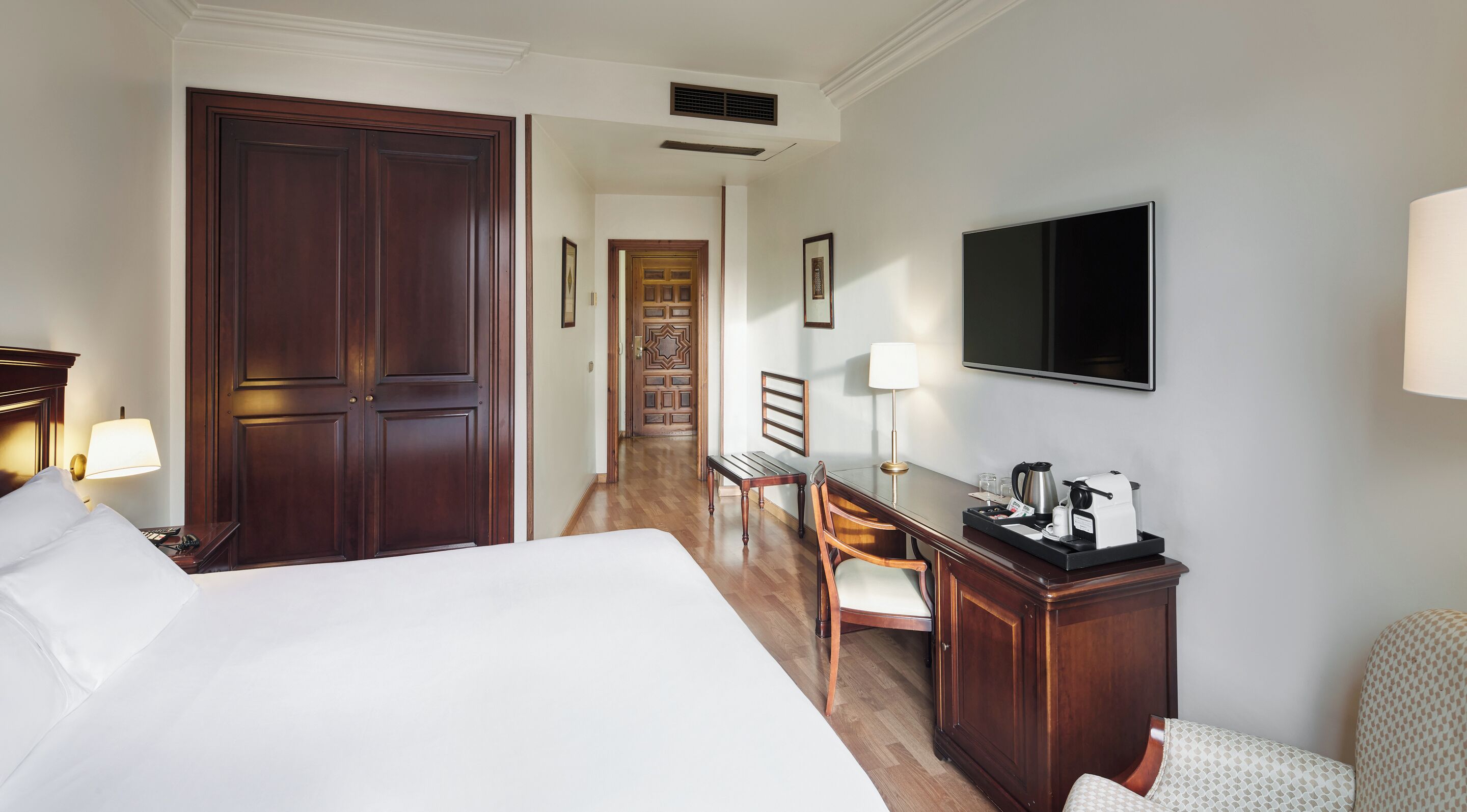 Habitación de hotel con una cama grande, un escritorio con cafetera, televisión y una puerta que da a un pasillo.