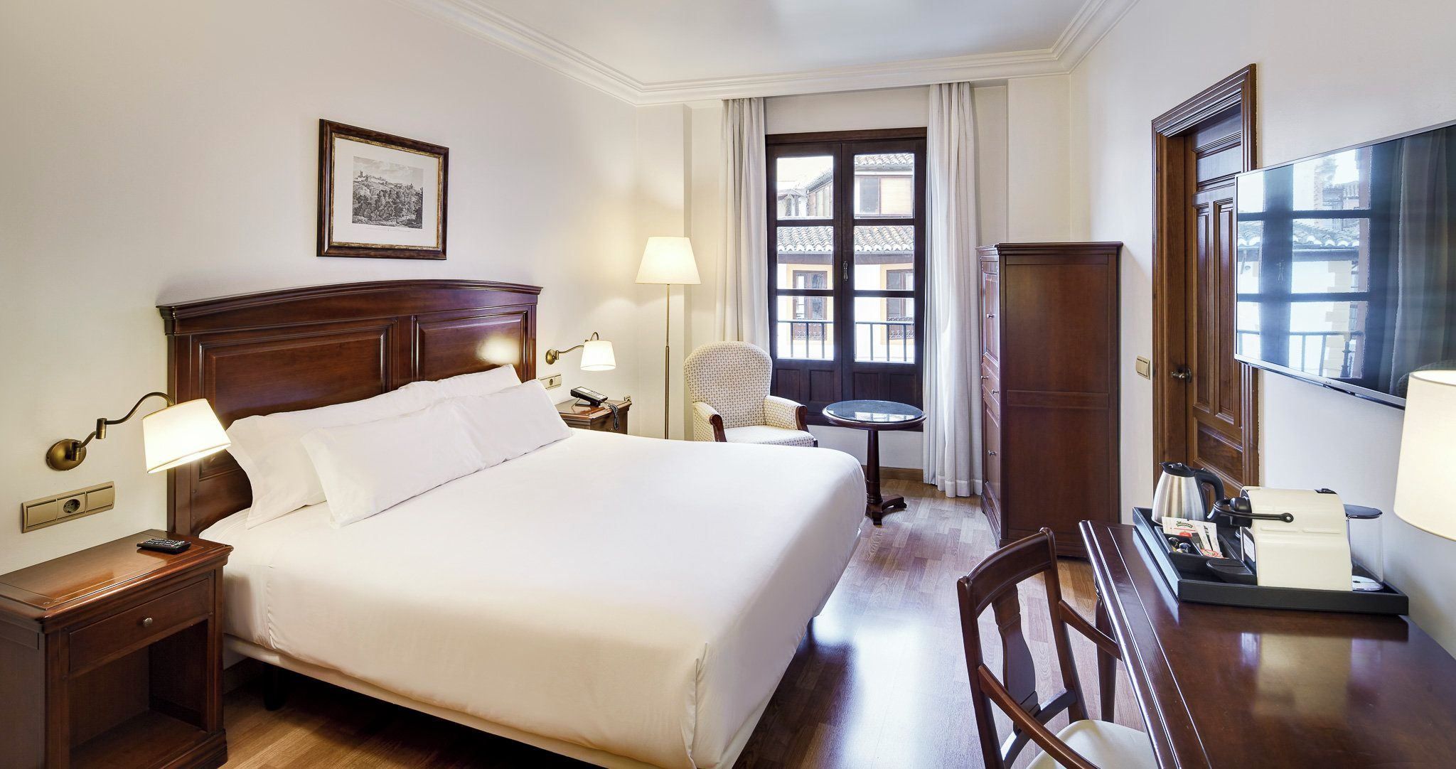 A hotel room with a large bed, bedside lamps, a chair, a desk with a lamp, a wardrobe, and a window with curtains.