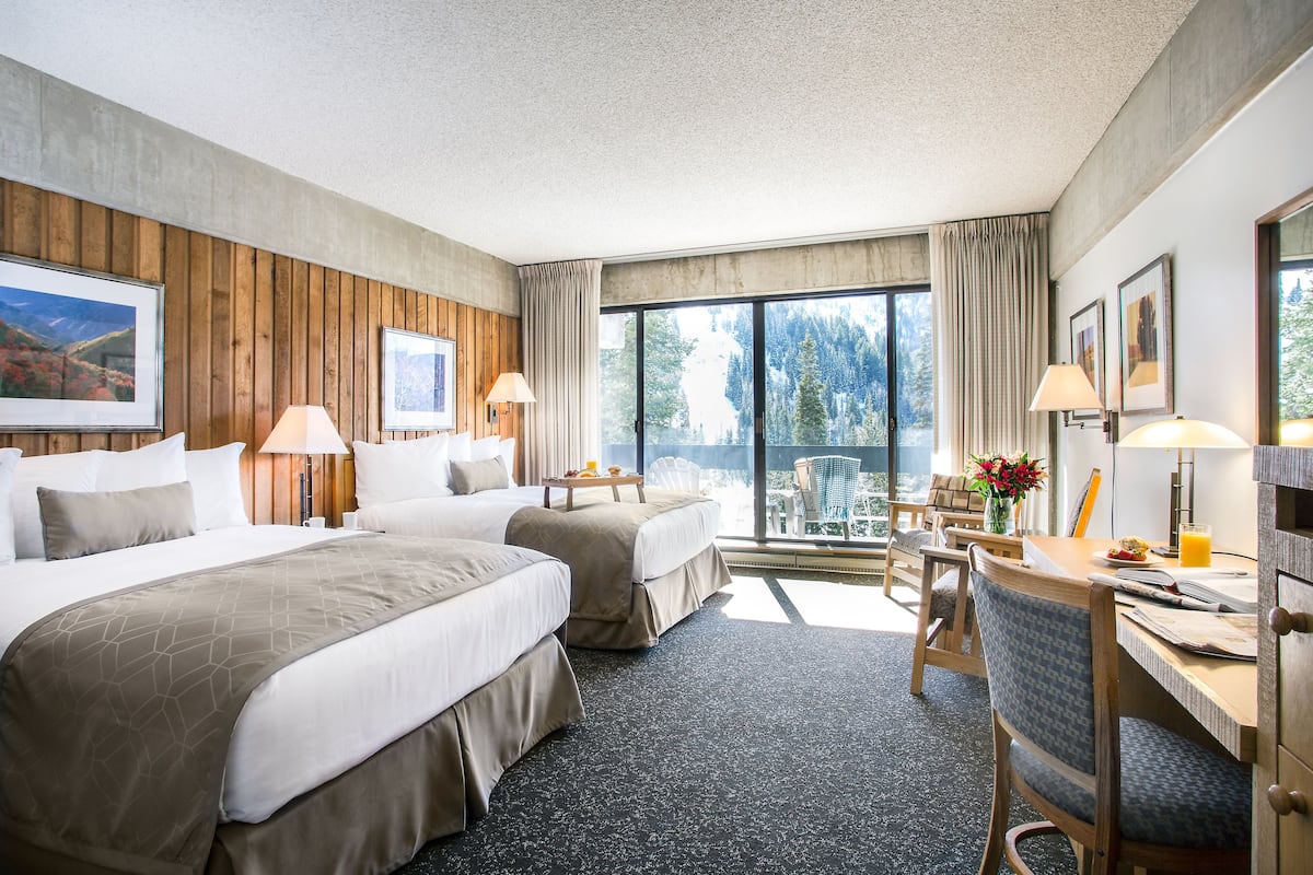 Ett hotellrum med två sängar, ett skrivbord och en stol. Det finns stora fönster med gardiner, utsikt över träd och en inramad tavla på väggen.