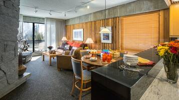 A modern hotel room with a dining area, a sofa, and a coffee table. There are two lamps on the dining table and a vase with flowers on the side table.