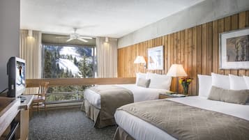 A hotel room with two beds, a desk, a chair, a TV, and a large window with a view of trees and mountains.