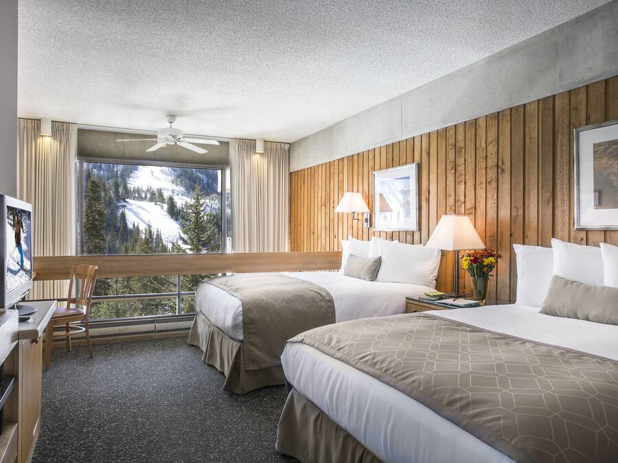 A hotel room with two beds, a desk, a chair, a TV, and a large window with a view of trees and mountains.