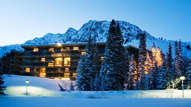 Un hôtel à plusieurs étages, doté d’un design moderne, entouré d’arbres couverts de neige et avec des montagnes en toile de fond.