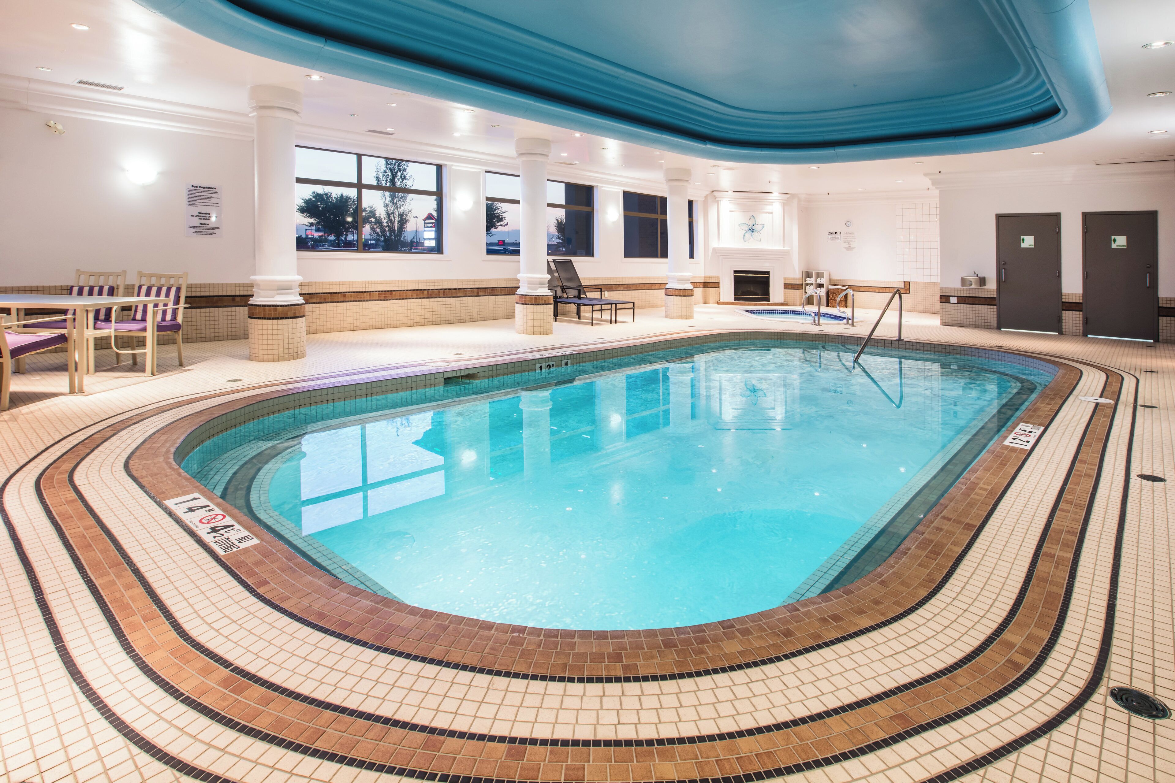 A large indoor swimming pool with clear water, surrounded by a tiled deck and a lounge area with chairs and tables.